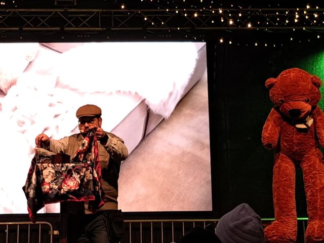 Fred Funke mit einem bunten Tuch auf einer Weihnachtsmarktbühne neben einem großen Teddybären